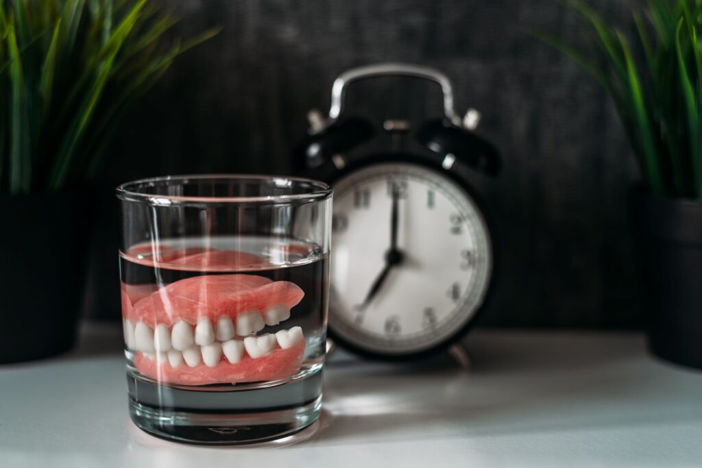 Full set of dentures in a clear glass on a desk next to an alarm clock